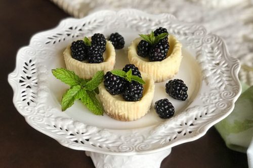 Three quark mini cheesecakes, each topped with blackberries and a sprig of mint, are placed on a decorative white plate with a lace-like pattern. The plate sits on a pedestal against a dark background, with part of a green cloth visible to the side.