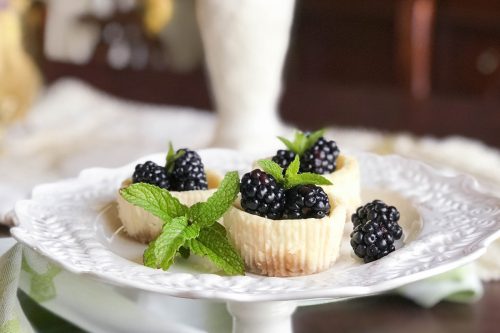 A decorative white cake stand holds three small quark mini cheesecakes topped with fresh blackberries and garnished with sprigs of mint. The background features a blurred, elegant setting with a white vase containing a floral arrangement.