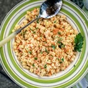 A plate of pasta salad with shredded carrots garnished with chopped herbs and paprika, accompanied by a serving spoon. The dish rests on an ornate metal table beside a green cloth napkin.
