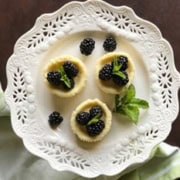 A delicate white lace-edged plate holds three quark mini cheesecakes topped with blackberries, with a mint sprig on the side. A light green cloth is draped next to the plate, and the background is a dark surface.