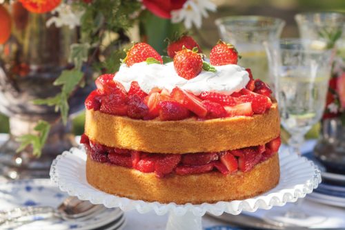 A two-layer strawberry shortcake topped with whipped cream and strawberries sits on a white cake stand. Perfect for a summer afternoon tea, bright red and orange flowers in a vase and assorted glassware adorn the background on a table with a blue and white tablecloth.