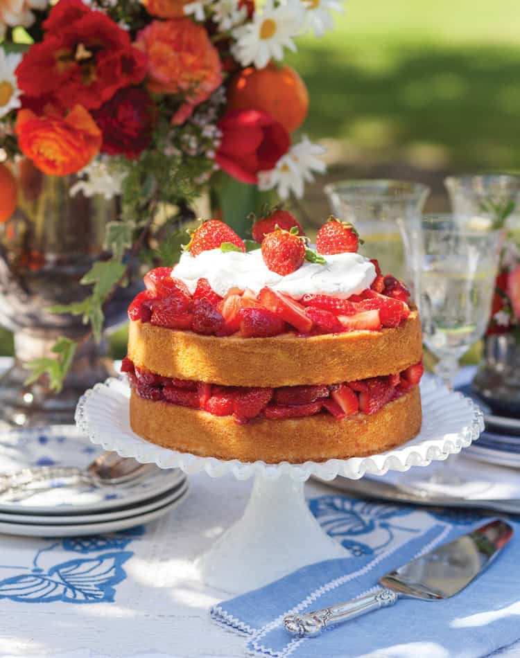 A two-layer strawberry shortcake topped with whipped cream and strawberries sits on a white cake stand. Perfect for a summer afternoon tea, bright red and orange flowers in a vase and assorted glassware adorn the background on a table with a blue and white tablecloth.