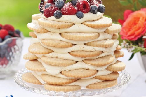 A layered stack of cookies filled with cream sits elegantly on a glass stand, topped with fresh strawberries, blueberries, and mint leaves. Perfect for a summer afternoon tea, the dessert rests on a white tablecloth adorned with floral decorations and blurred greenery in the background.