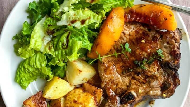 A white plate with skillet pork chops and peaches, roasted potatoes, leafy green salad, and peach slices, with a fork on the side.