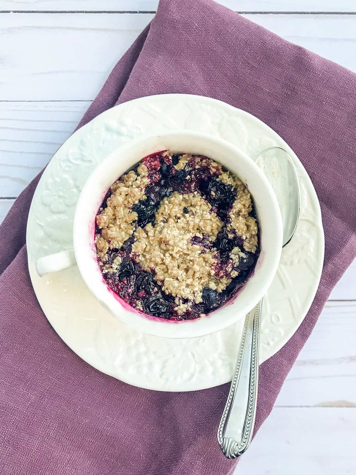 A white cup with blueberry crisp in a mug, featuring baked oatmeal and berries, sits on a white plate with a silver spoon, placed on a purple napkin atop a white wooden surface-perfect for making quickly in the microwave.