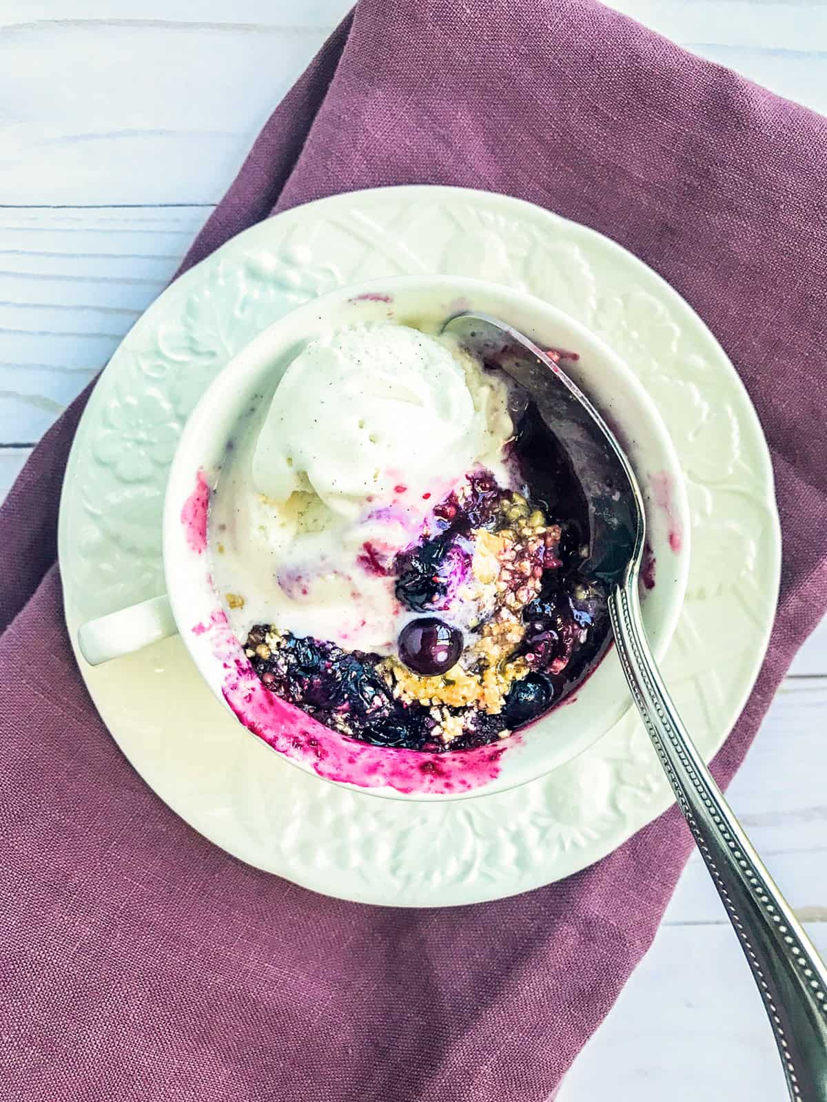 A cup filled with Blueberry Crisp in a Mug, topped with a scoop of vanilla ice cream, sits on a white saucer with a spoon, placed on a folded mauve napkin-perfect for a quick microwave treat.