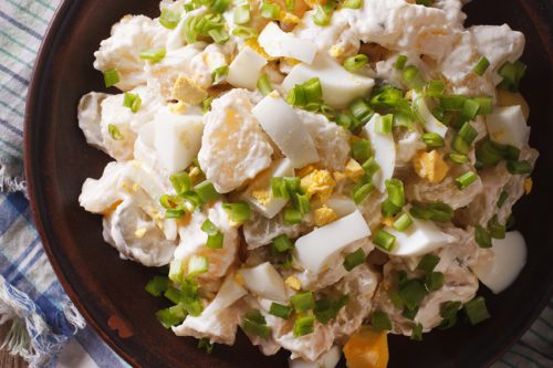A plate of creamy potato salad recipe topped with chopped hard-boiled eggs and green onions, set on a rustic wooden table with a plaid cloth underneath.
