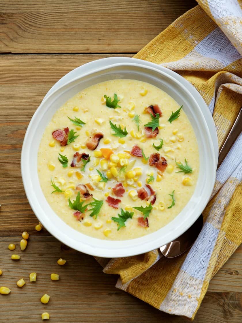 A bowl of creamy corn chowder, perfect for dinner, is topped with crispy bacon bits and fresh parsley on a wooden table. A yellow and white checkered napkin with a spoon rests beside the bowl. Scattered corn kernels complete the rustic presentation.