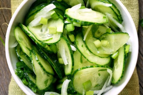 A white bowl filled with cucumber salad-sliced cucumbers, onions, and chopped green herbs-rests on a light green textured cloth atop a wooden surface.