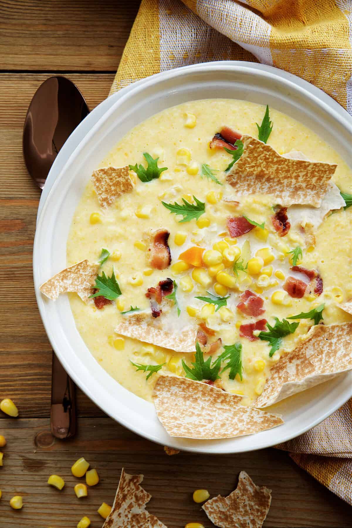 A bowl of creamy Corn Chowder topped with bacon bits, parsley, corn kernels, and broken pieces of flatbread sits on a wooden surface beside a brown spoon.