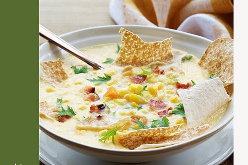 A bowl of hearty Corn Chowder with bacon, vegetables, and crackers on top, served on a plate with a spoon beside a napkin.