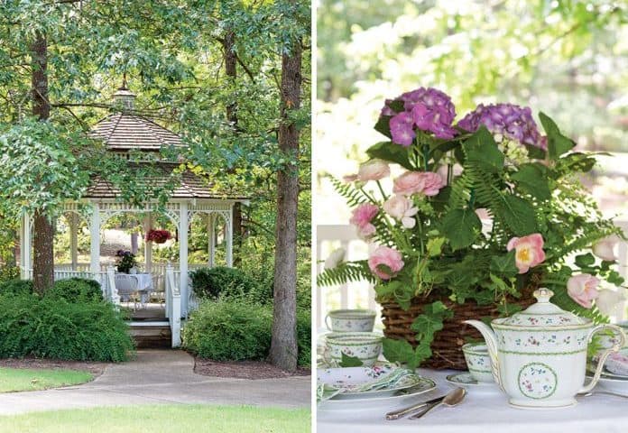 A white gazebo surrounded by lush greenery on the left. On the right, a table set for a summer afternoon tea with floral-patterned china, a teapot, and a basket of pink and purple flowers with ferns. A peaceful garden setting.