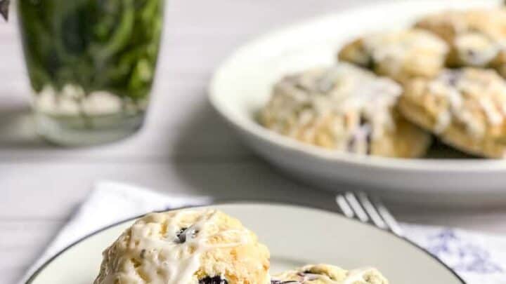 Two blueberry scones with white icing sit on a plate, while more blueberry scones are arranged on a platter beside a vase of flowers in the background.