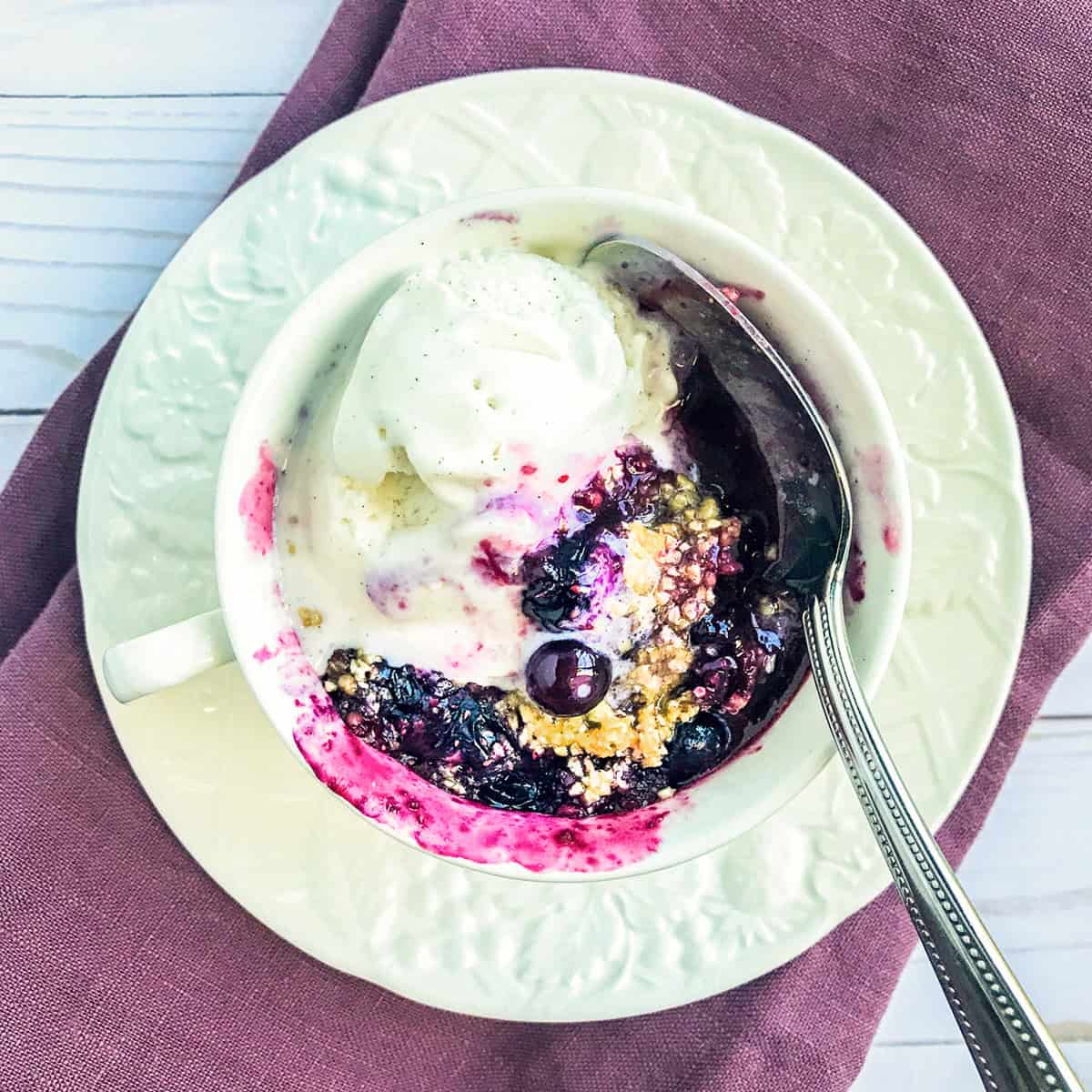 A delicious Blueberry Crisp in a Mug with a scoop of vanilla ice cream, served on a white plate with a spoon, sits atop a purple cloth on a light surface—perfect for enjoying straight from the microwave.