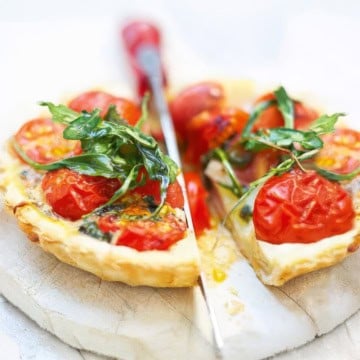 A savory tomato tart with cherry tomatoes and fresh arugula is cut into slices on a white surface, with a knife placed between the pieces.