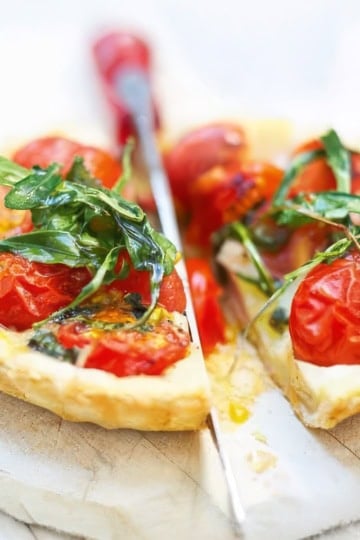 A savory tomato tart with cherry tomatoes and fresh arugula is cut into slices on a white surface, with a knife placed between the pieces.