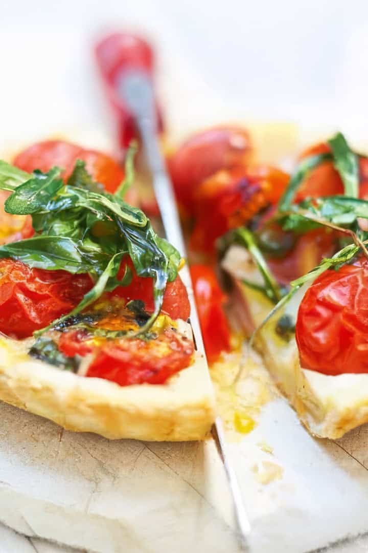 A savory tomato tart with cherry tomatoes and fresh arugula is cut into slices on a white surface, with a knife placed between the pieces.