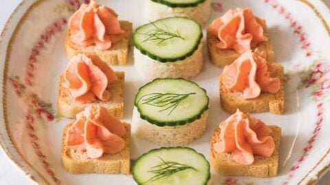 Plate of afternoon tea savory canapés with round cucumber and dill sandwiches and rectangular toasts topped with a swirl of pink spread, arranged neatly on a floral-patterned dish.