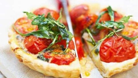A slice of savory pizza being cut, topped with cherry tomatoes, arugula, and melted cheese on a thin crust. The vibrant colors and fresh ingredients are highlighted like an afternoon tea spread. The pizza rests on a light cutting board.
