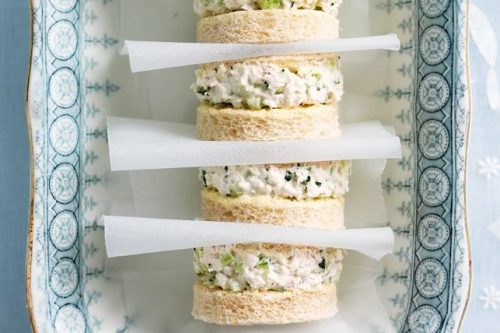Stacked sandwiches on a decorative blue and white platter evoke the charm of a summer afternoon tea. Round slices of white bread cradle creamy chicken salad with celery and herbs, each separated by strips of white parchment paper for a delightful touch.