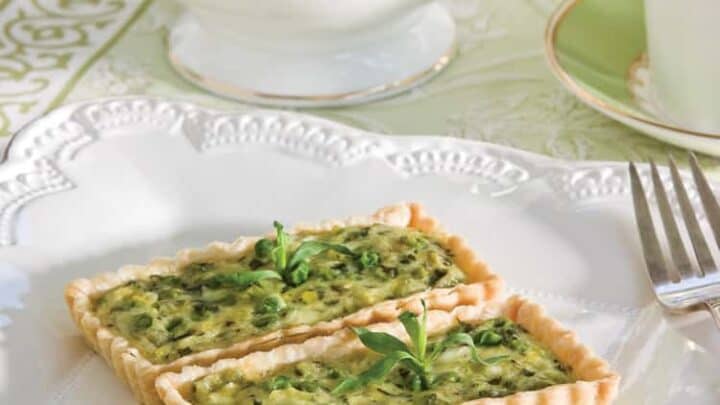 Two rectangular slices of savory vegetable quiche are served on a white plate with a fork, perfect for enjoying during afternoon tea, with a charming tea set in the background.