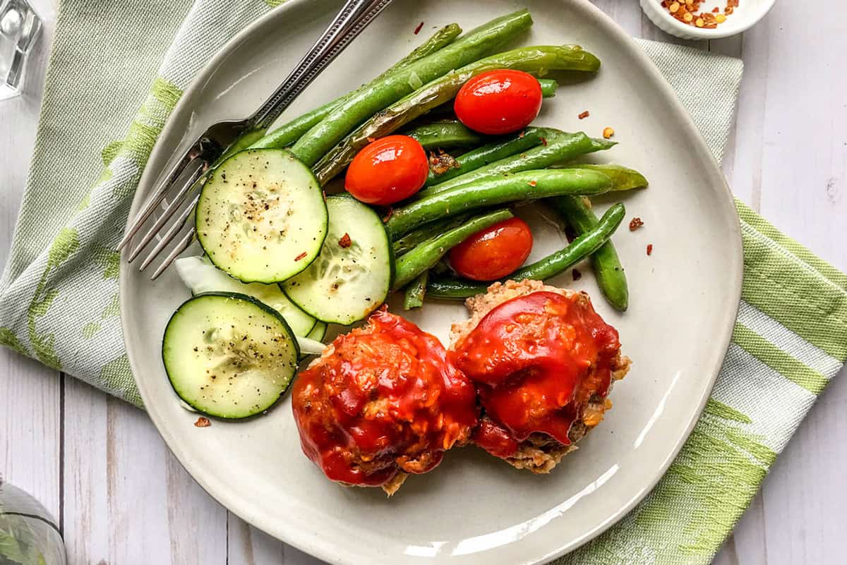 A plate with two Mini Turkey Meatloaves topped with tomato sauce, green beans, grape tomatoes, and cucumber slices, served with a fork and sprinkled seasonings on a green-striped napkin.