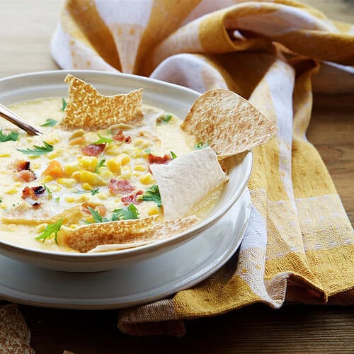 A bowl of creamy corn chowder garnished with bacon bits, herbs, corn kernels, and tortilla chips, with a spoon and a yellow-checkered cloth beside it.