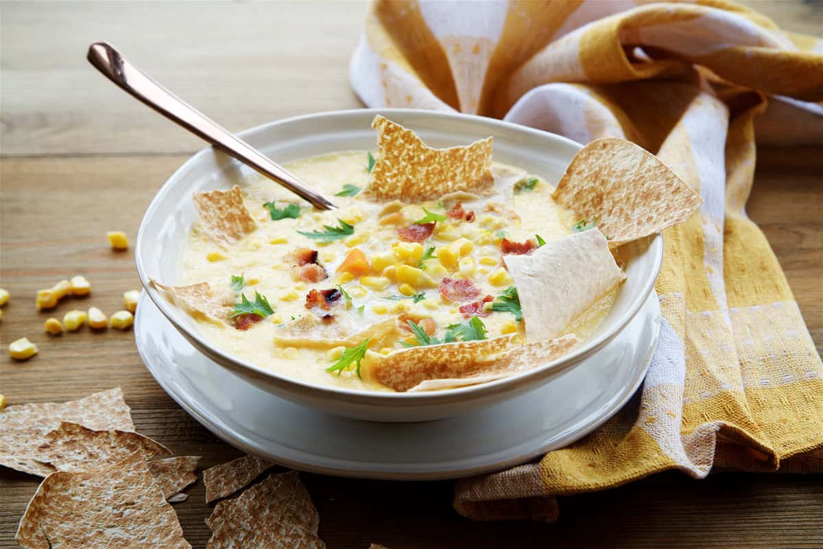 A bowl of creamy corn chowder garnished with bacon bits, herbs, corn kernels, and tortilla chips, with a spoon and a yellow-checkered cloth beside it.