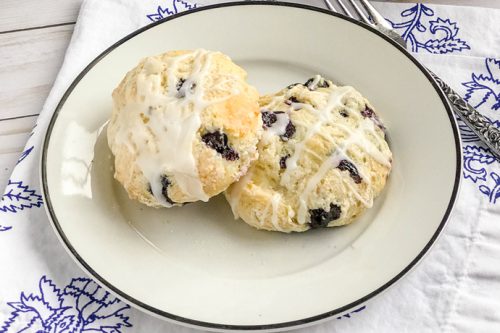 Two blueberry scones with white icing drizzle sit on a white plate with a black rim, placed atop a blue and white patterned napkin-perfect for blueberry scone lovers seeking a delightful treat.