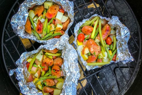 Opened sausage and vegetable foil packets on a grill.