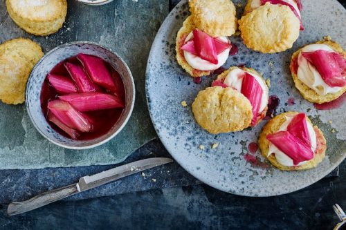 A blue plate showcases scones adorned with cream and vibrant pink rhubarb, evoking a perfect summer afternoon tea. A nearby bowl brims with extra rhubarb beside a knife on the textured surface.