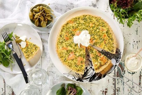 A quiche garnished with a dollop of cream sits on a white plate, surrounded by a slice on another plate, mixed greens, and capers-perfect for a summer afternoon tea. Two glasses sit nearby on a rustic white table.