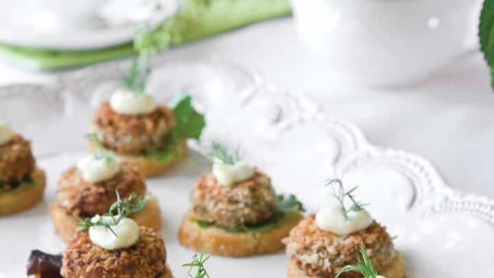 A platter of bread rounds topped with leafy greens, breaded patties, and a dollop of creamy sauce makes a perfect afternoon tea savory, served on a white tray with a floral centerpiece and teacup in the background.