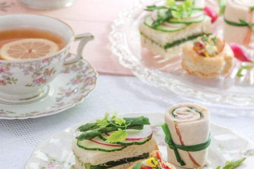 A delicate summer afternoon tea setting with a floral teacup and saucer filled with tea. Plates hold elegant finger sandwiches, wraps, and pastry bites. A small radish and sprig of greens garnish the dishes, set on a lace tablecloth.
