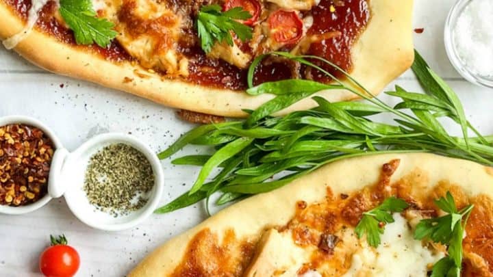 Two homemade bbq chicken pizzas topped with melted cheese, cherry tomatoes, and parsley are displayed on a white surface, with small bowls of spices nearby.
