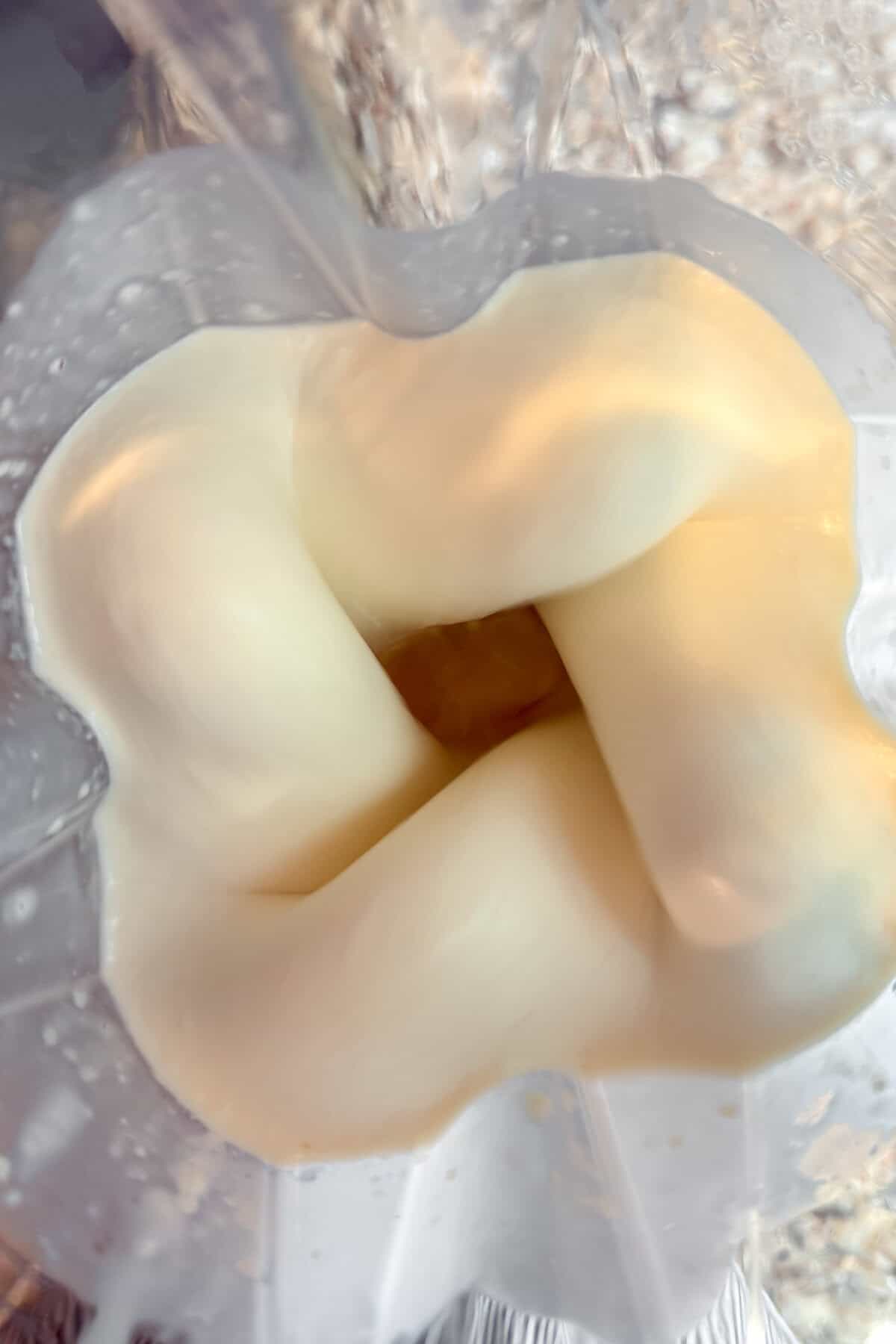 Close-up view from above of a blender mixing a creamy, light-colored pineapple smoothie, creating a swirling vortex in the center of the blender jar.