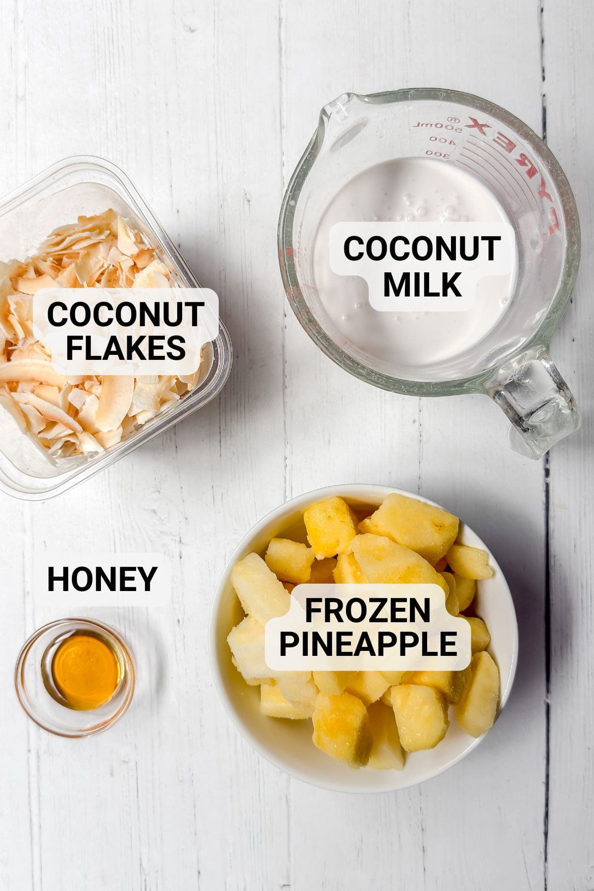 Flat lay of ingredients for a pineapple smoothie on a white surface: coconut flakes, coconut milk in a glass measuring cup, frozen pineapple chunks in a bowl, and honey in a small glass dish.