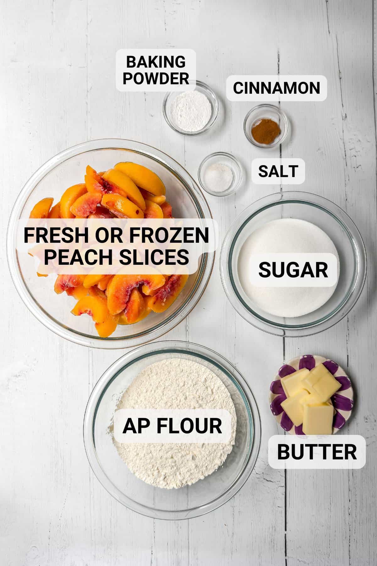 Top-down view of labeled baking ingredients for slow cooker peach cobbler in bowls: fresh or frozen peach slices, all-purpose flour, sugar, butter, baking powder, cinnamon, and salt on a white wooden surface.