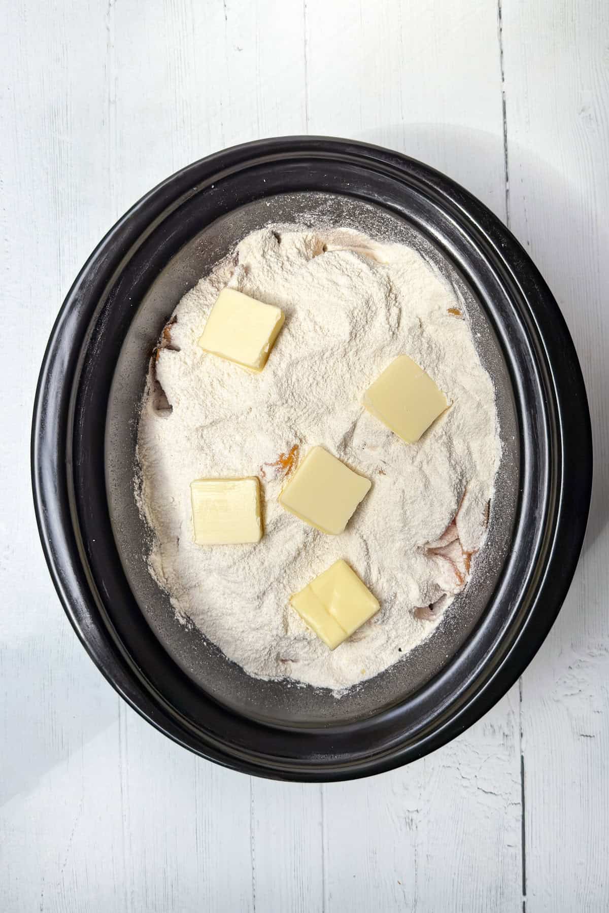 A black slow cooker filled with flour and several slices of butter on top, ready to be transformed into a delicious slow cooker peach cobbler, sits on a white wooden surface.