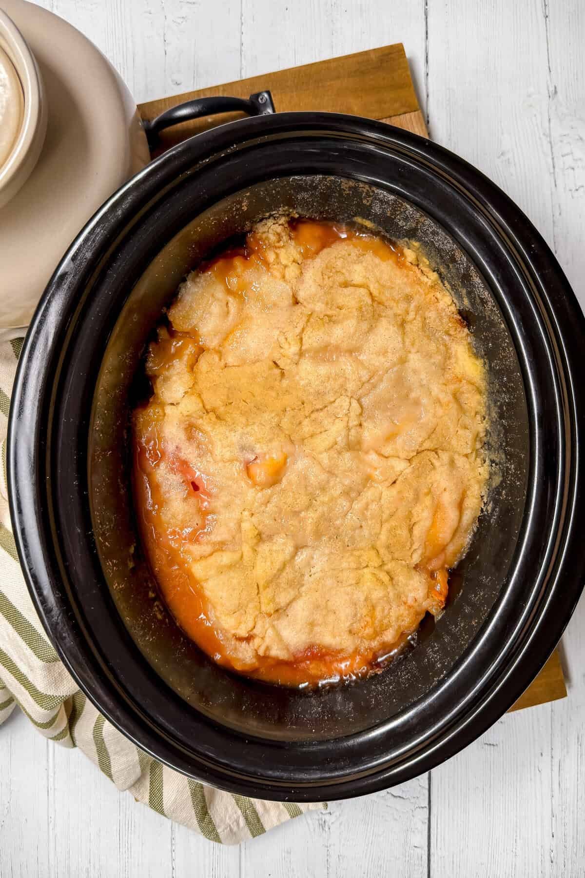 A slow cooker peach cobbler with a golden-brown crust and bubbling peach filling sits on a wooden board, with a striped cloth partially visible underneath.