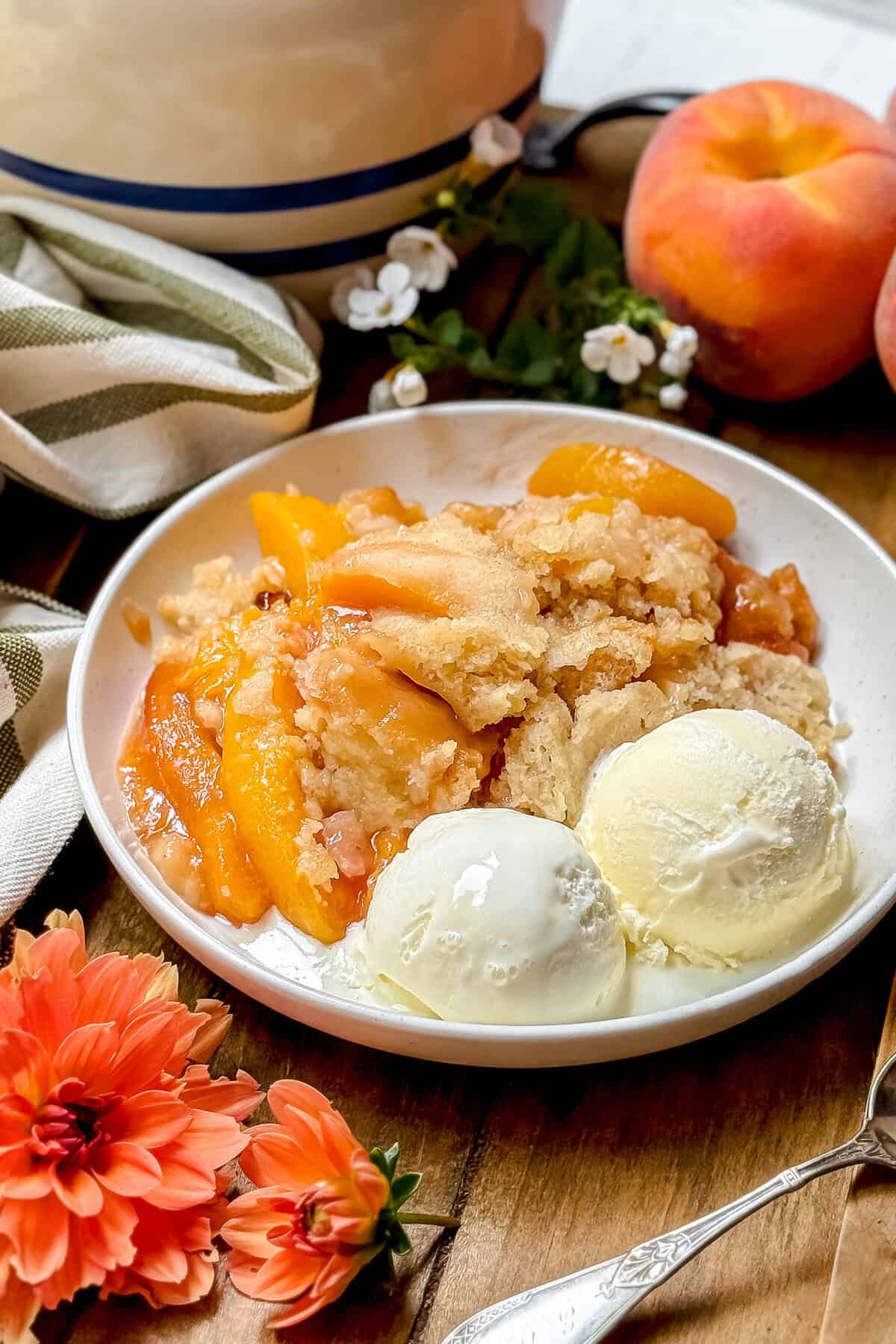 A plate with slow cooker peach cobbler and two scoops of vanilla ice cream, surrounded by fresh peaches, flowers, a striped towel, and a baking dish in the background.
