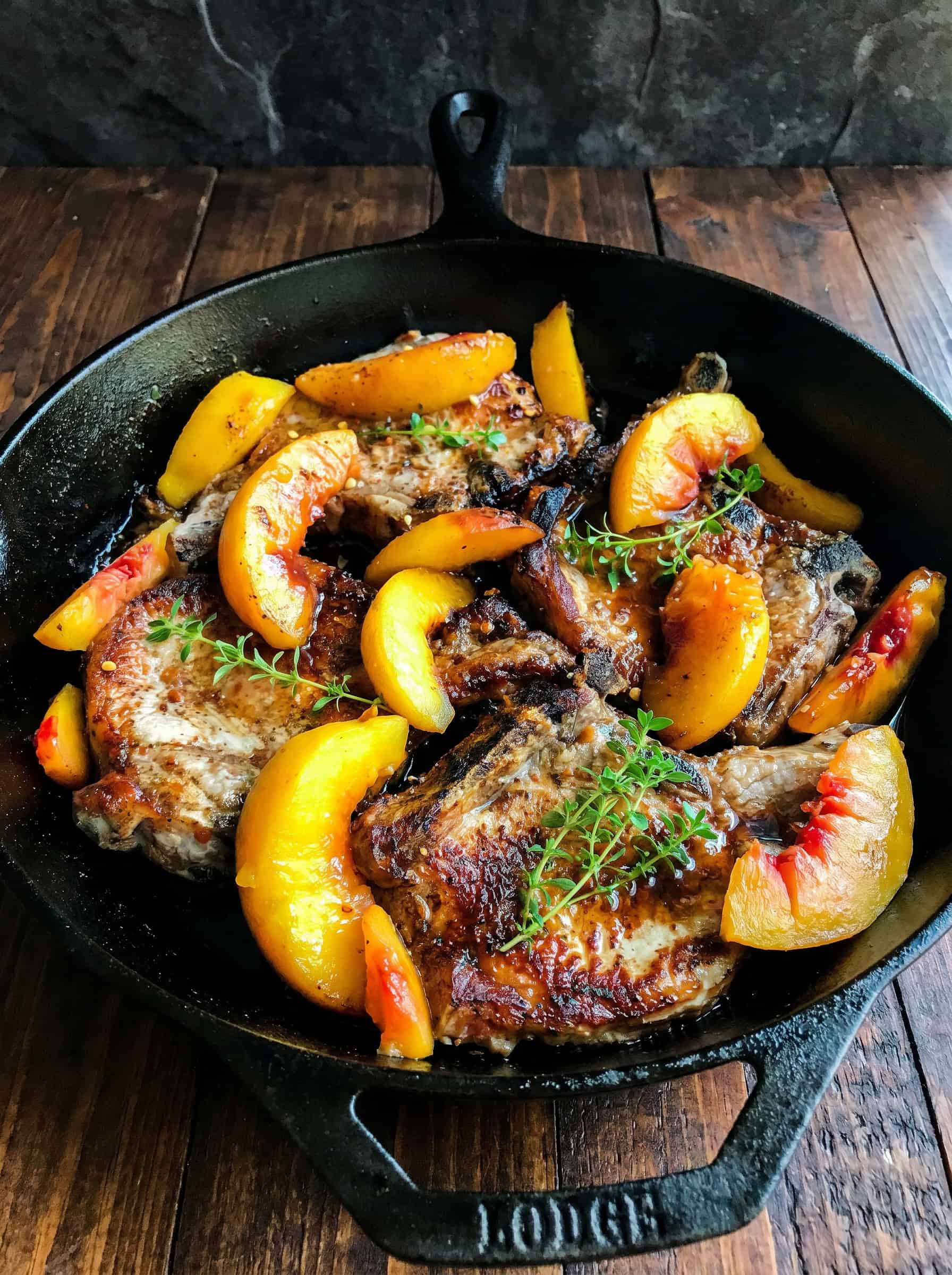 Skillet pork chops and peaches cooked in a cast iron pan, garnished with fresh thyme, served on a rustic wooden surface.