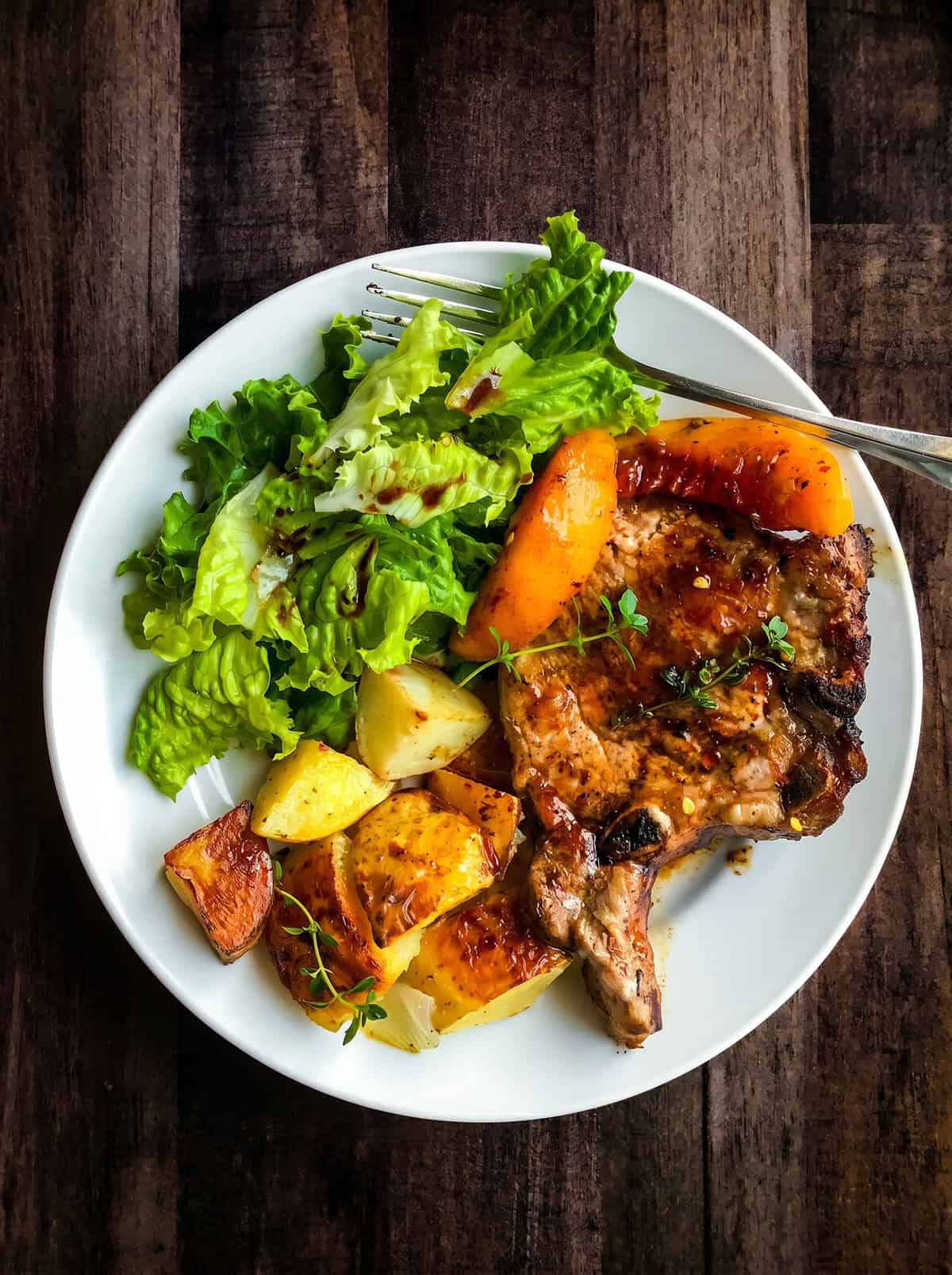 A white plate with skillet pork chops and peaches, roasted potatoes, lettuce salad, and orange bell pepper sits on a wooden table with a fork.