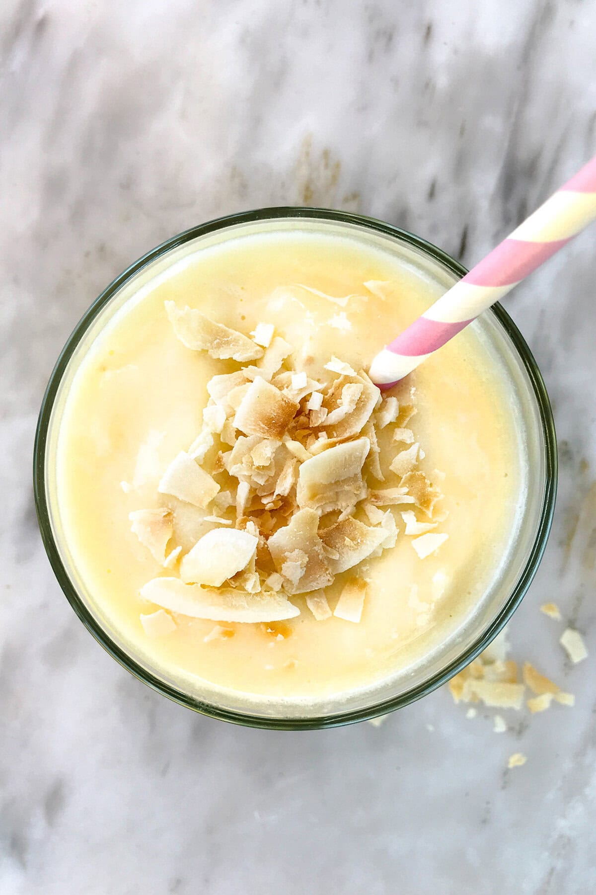 A top-down view of a creamy yellow pineapple smoothie in a glass, topped with coconut flakes, with a pink and yellow striped straw on a light marble surface.