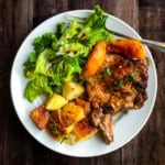 A white plate with skillet pork chops and peaches, roasted potatoes, and a green leafy salad with dressing sits on a dark wooden table beside a fork.