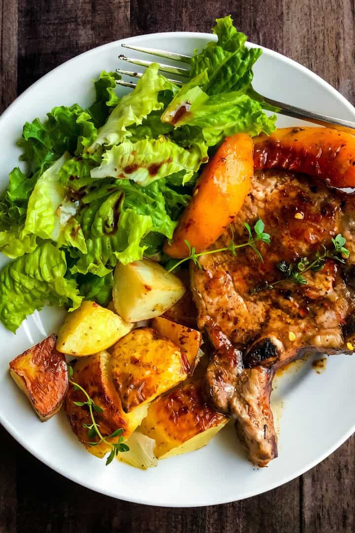 A white plate with skillet pork chops and peaches, roasted potatoes, and a green leafy salad with dressing sits on a dark wooden table beside a fork.