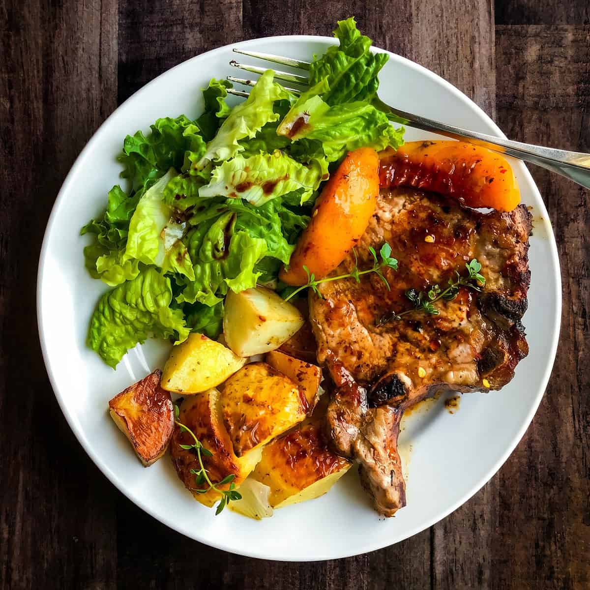 A white plate with skillet pork chops and peaches, roasted potatoes, and a green leafy salad with dressing sits on a dark wooden table beside a fork.