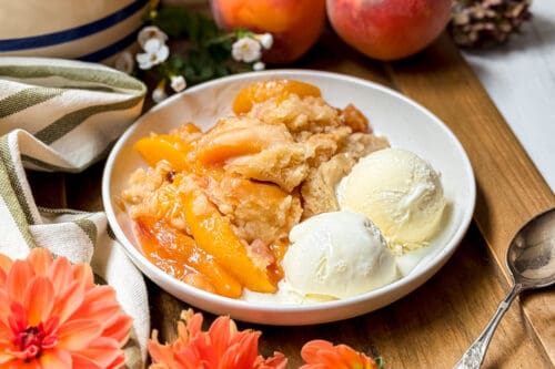 A plate of slow cooker peach cobbler with golden baked topping, served with two scoops of vanilla ice cream. Fresh peaches and flowers decorate the wooden table beside a striped cloth and a spoon.