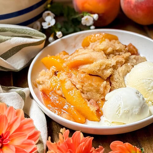 A plate of slow cooker peach cobbler with golden baked topping, served with two scoops of vanilla ice cream. Fresh peaches and flowers decorate the wooden table beside a striped cloth and a spoon.