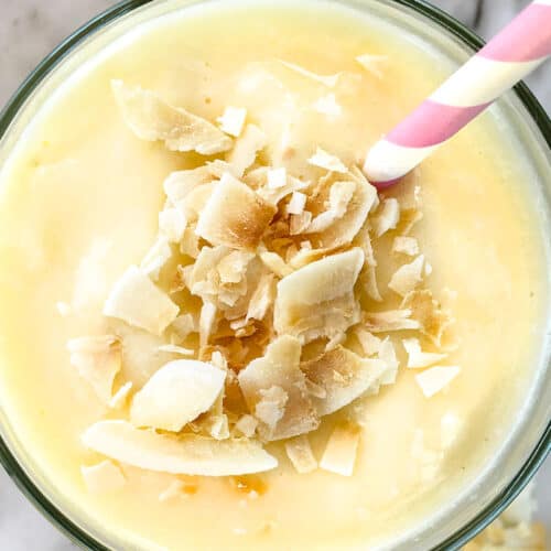 A creamy yellow pineapple smoothie topped with toasted coconut flakes, served in a glass with a pink-and-white striped straw on a light, marble surface.